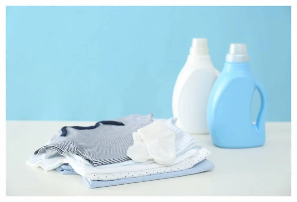 Pile de vêtements de bébé bleus, gris et blancs sur un comptoir avec une bouteille de détergent à l'arrière-plan.