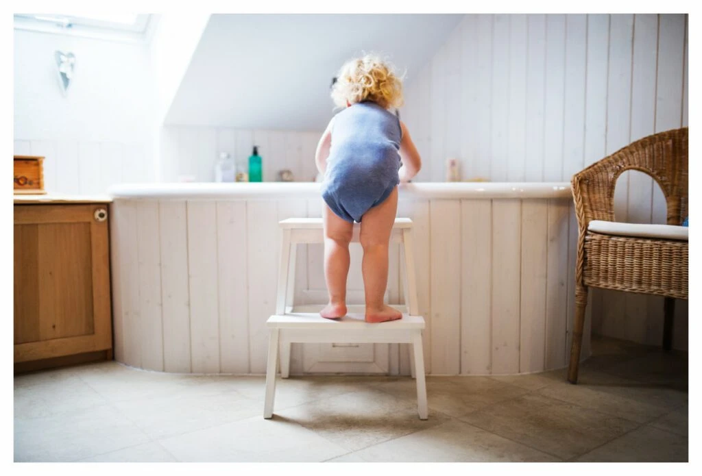 Ein Kleinkind in einem blauen Strampler steht auf einer Treppe (von IKEA) und schaut in eine Badewanne. Auf dem Regal hinter der Wanne stehen eine Reihe von Badezusätzen, aber seltsamerweise kein Badespielzeug. 