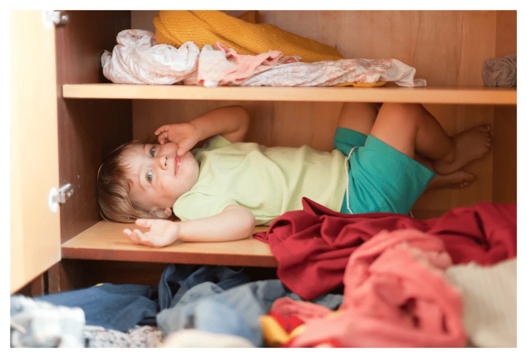Enfant effronté se cachant sur l'étagère de son armoire à vêtements. Les vêtements sont par terre au premier plan !