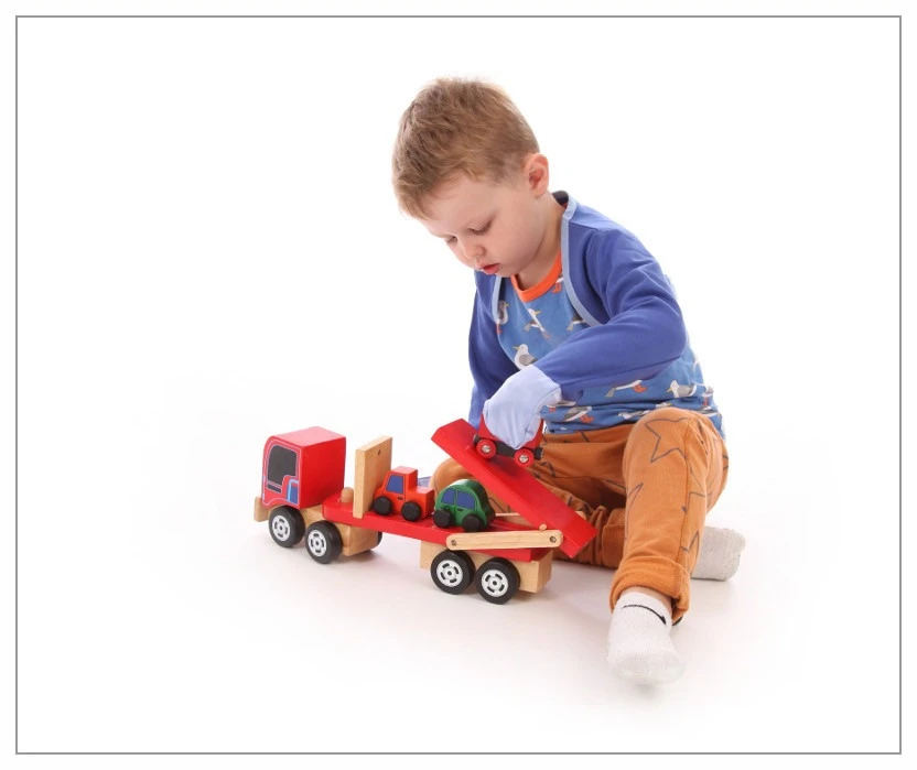 Kleiner Junge mit Kratzhandschuhen, der mit großen Holzautos und einem Autotransporter-LKW spielt. Die Kratzhandschuhe haben offensichtlich nicht in den Weg seines Spiels zu bekommen.