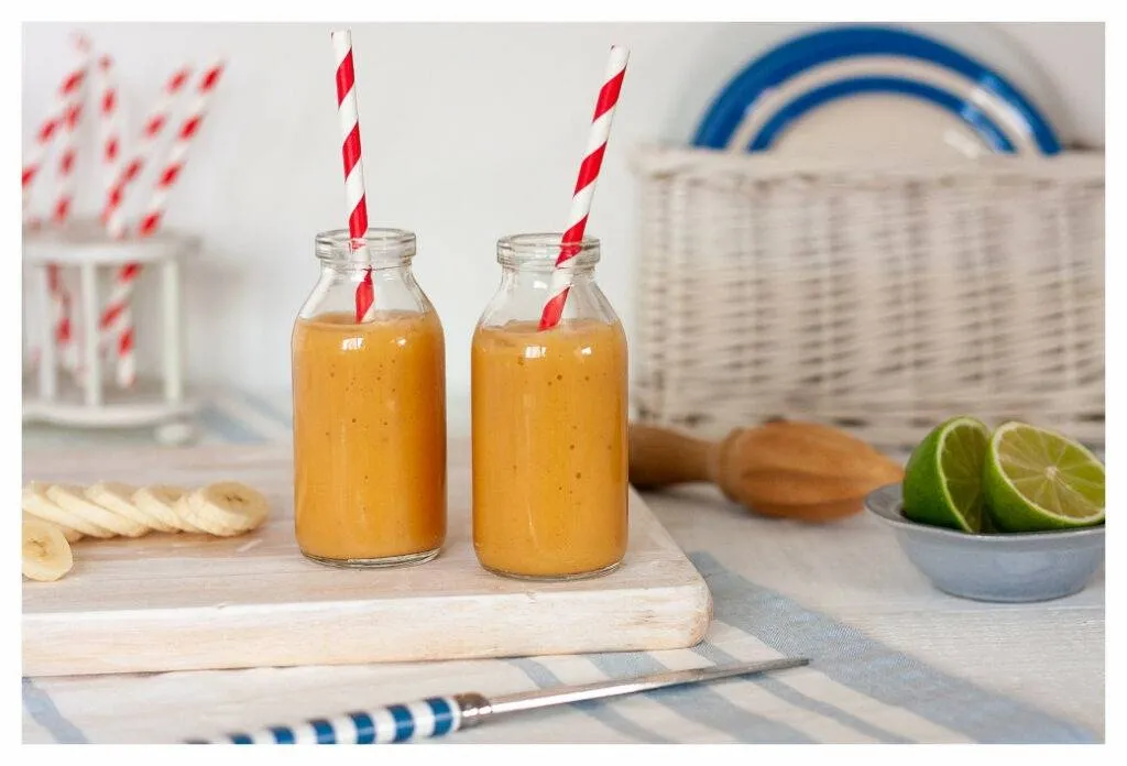 Ekzemfreundliche Papaya- und Bananen-Smoothies, serviert in Mini-Milchflaschen mit rot-weiß gestreiften Papierstrohhalmen. Der orangefarbene Smoothie kontrastiert mit dem blau-weißen Hintergrund.